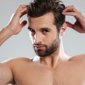 Close up portrait of a charming naked bearded man looking at camera and touching his hair isolated over white background